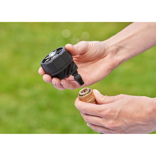 Close-up of two hands holding a BLACK+DECKER hose nozzle and a brass connector, with a grassy background.