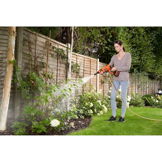 A person using a BLACK+DECKER portable water sprayer to water plants in a garden, standing on grass near a wooden fence.