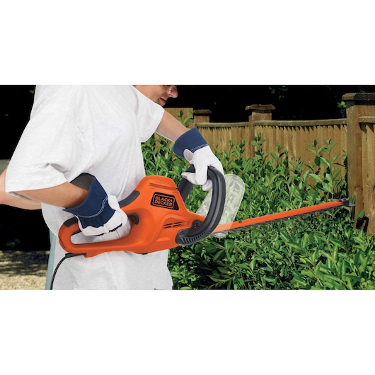 Photo of BLACK+DECKER GT7030 hedge trimmer in use, orange electric tool cutting green hedge near wooden fence.