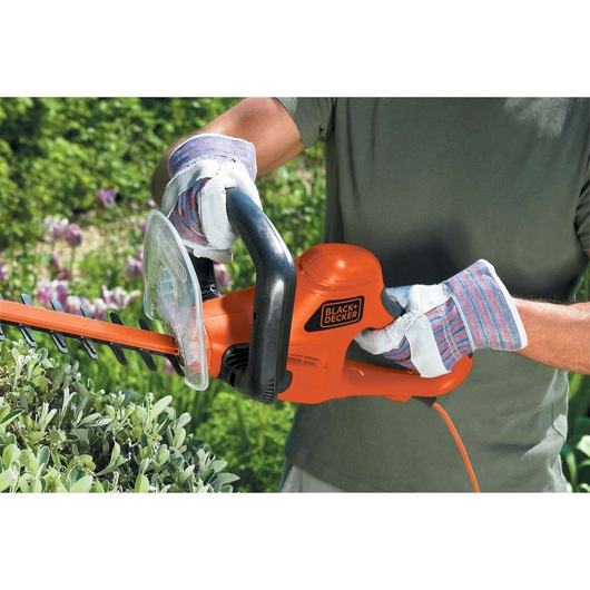 BLACK+DECKER GT7030 electric hedge trimmer in use, orange body, user wearing gloves, trimming green bushes outdoors.