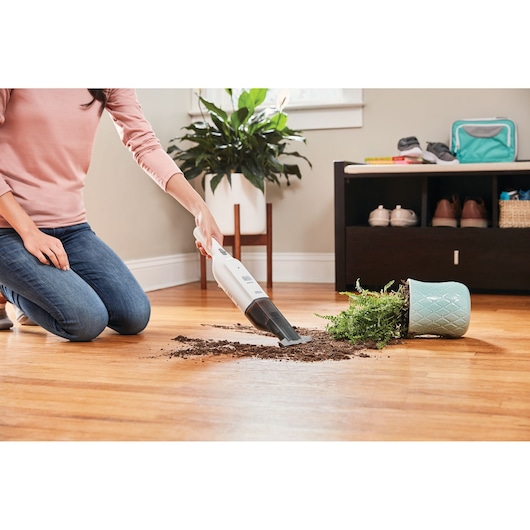 BLACKDECKER HLVC315B11 handheld vacuum cleaning spilled soil from a plant on a wooden floor, indoor setting.