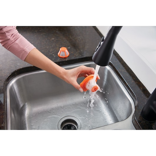 Photo of a hand rinsing BLACK+DECKER HLVC315B11 vacuum filter under a kitchen faucet over a stainless steel sink.