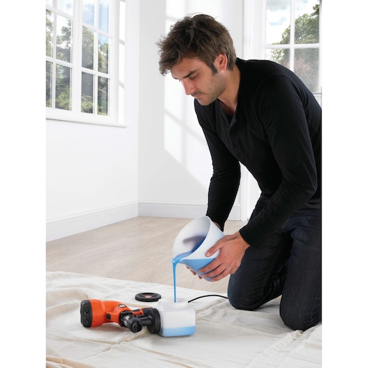Photo of a person pouring blue paint into BLACK+DECKER HVLP200 paint sprayer container indoors on a white sheet.