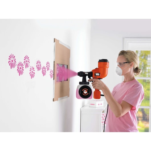 BLACK+DECKER HVLP200 paint sprayer being used to paint pink flower patterns on a white wall with a stencil.