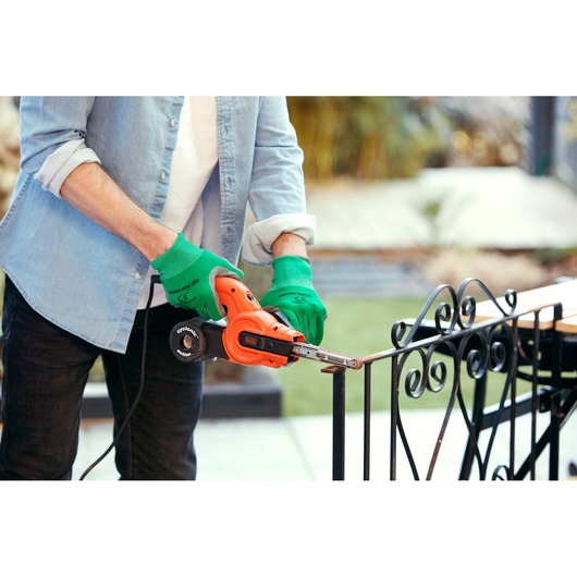 Person wearing green gloves using an orange BLACK+DECKER KA900E power tool to sand the metal railing outdoors.