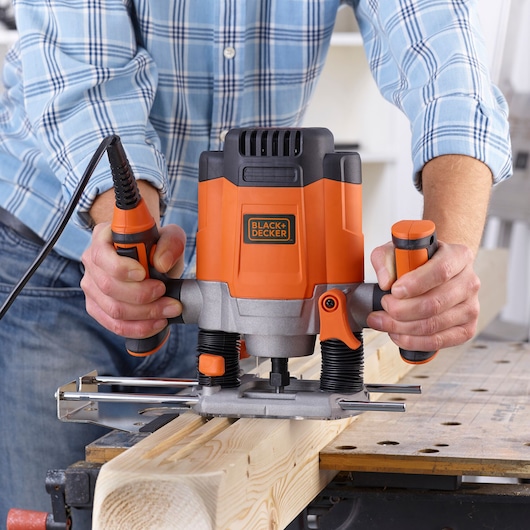A person using a BLACK+DECKER KW1200EKA electric wood router to shape a piece of wood on a workbench. The device is orange and black and is being held firmly by both hands.