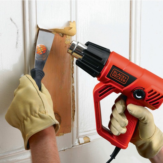 Photo of BLACKDECKER KX2001K heat gun used for paint removal on wooden door, held by gloved hands.