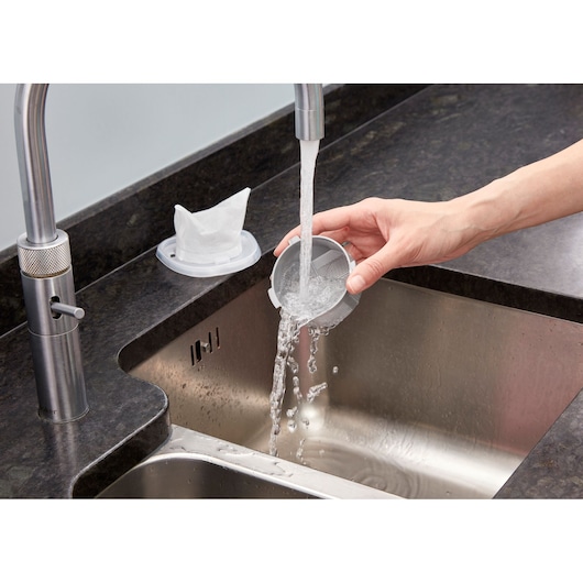 Photo of a hand rinsing BLACKDECKER NVC115JL vacuum filter under a kitchen faucet, sink and counter visible.