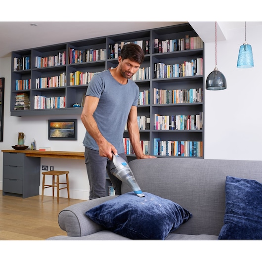 BLACKDECKER NVC215WA handheld vacuum cleaning blue cushion on grey sofa in modern living room.