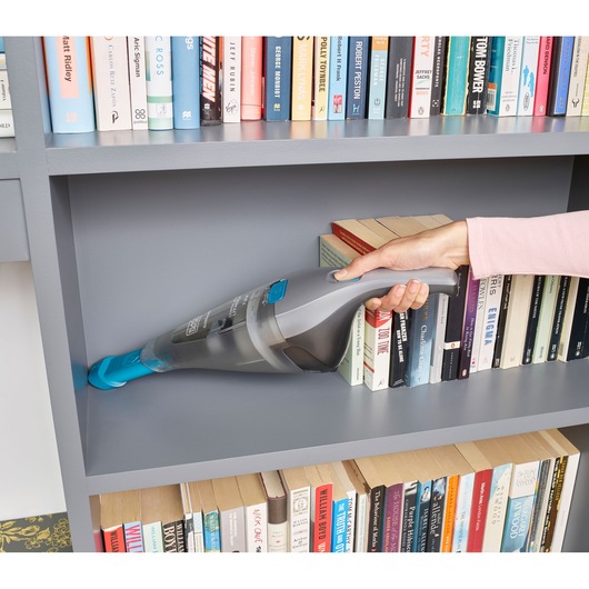 BLACK+DECKER NVC215WA handheld vacuum with blue nozzle cleaning a gray bookshelf, books visible in background.