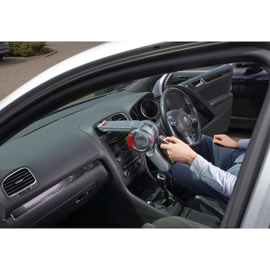 A person using a BLACK+DECKER handheld vacuum cleaner (model PV1200AV) to clean the dashboard inside a car.