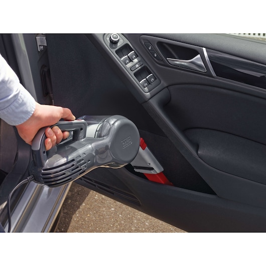 A person is using a BLACK+DECKER handheld vacuum cleaner to clean the interior of a car door.