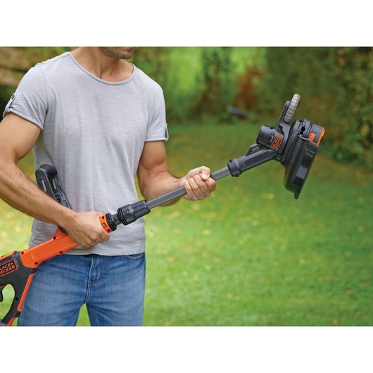 A person outdoors holding a BLACK+DECKER battery-powered grass trimmer with an adjustable shaft.