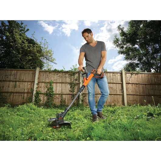 A person using a BLACK+DECKER STC1840EPC cordless grass trimmer on a lawn, with a wooden fence and trees in the background.