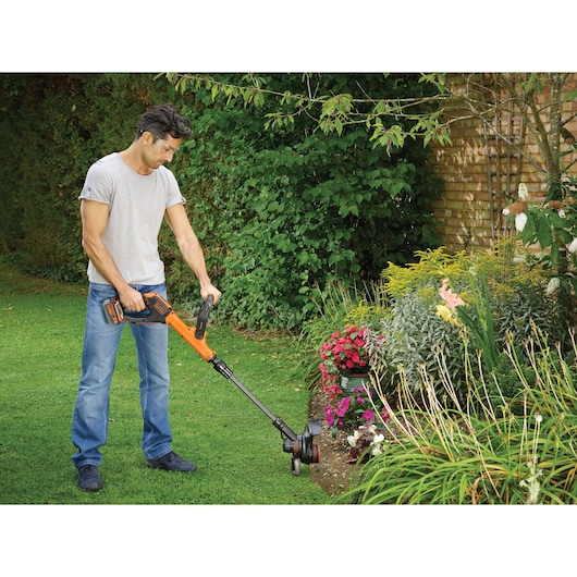 A person is using a BLACK+DECKER cordless grass trimmer, model STC1840EPC, to trim the edge of a garden lawn next to a flower bed. The tool is orange and black, and the person is working outdoors in a garden.