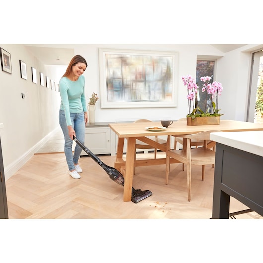 BLACK+DECKER SVB620JW cordless stick vacuum cleaning under wooden dining table, crumbs visible on light wood floor.